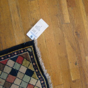 Multicolored diamond pattern rug corner with navy border and fringe sits on worn wooden planks.