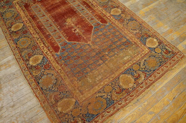 Low-pile rug with a rust red niche field and elaborate gold and blue floral borders on wood planks.