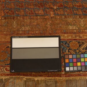 Worn, low-pile rug in rust, tan, and blue patterns rests on wood with color calibration charts.