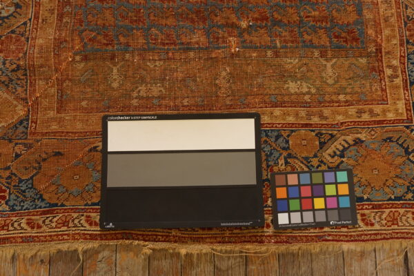 Worn, low-pile rug in rust, tan, and blue patterns rests on wood with color calibration charts.