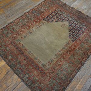 A faded low-pile rug with a khaki central field, navy accents, and red floral borders rests on rustic wood flooring.