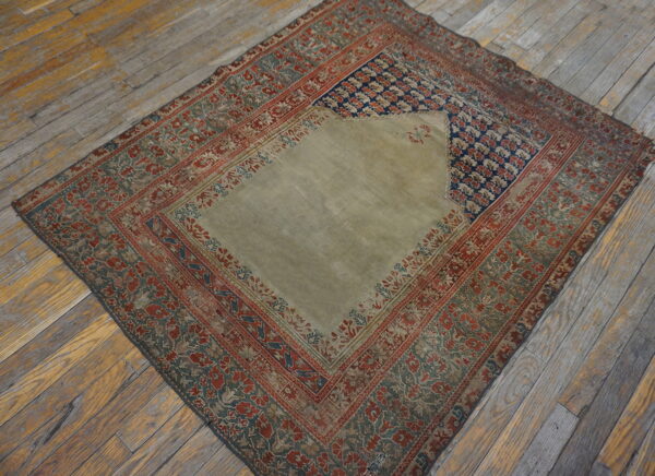 A faded low-pile rug with a khaki central field, navy accents, and red floral borders rests on rustic wood flooring.