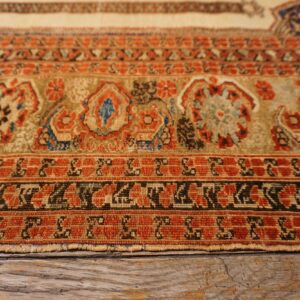 Rust, tan, and blue stylized patterns on a low pile rug edge, resting on cracked wooden flooring.