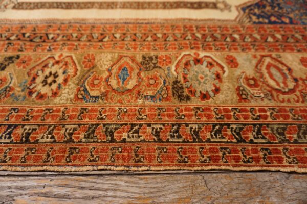 Rust, tan, and blue stylized patterns on a low pile rug edge, resting on cracked wooden flooring.