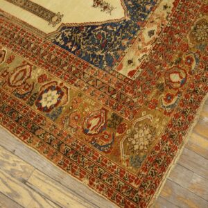 Traditional low-pile rug corner in gold, rust, and navy patterns rests on light, worn wooden floorboards.