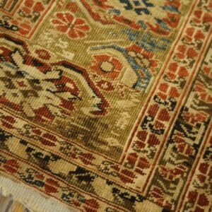 Worn, low-pile rug corner featuring traditional floral and geometric patterns in rust, gold, and blue colors on wood flooring.