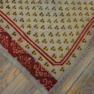 Close-up of a rug corner featuring pale yellow geometric motifs, red borders, and fringe over rustic wood flooring.