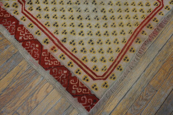 Close-up of a rug corner featuring pale yellow geometric motifs, red borders, and fringe over rustic wood flooring.