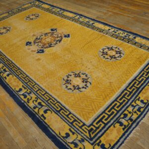 Gold and blue rug with medallions and a fret border rests on worn wood plank flooring.