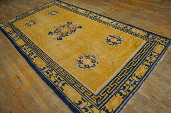 Gold and blue rug with medallions and a fret border rests on worn wood plank flooring.
