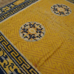 Gold rug with chevron texture and blue and white medallions bordered by a dark blue Greek key design.