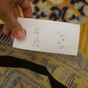 A hand holds a white tag over the tightly woven striped edge of a yellow and blue patterned rug.