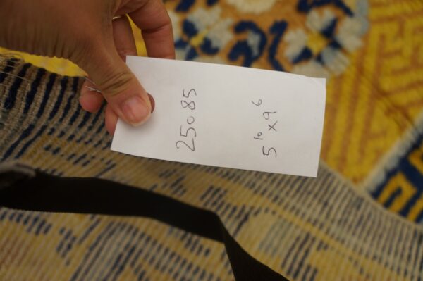 A hand holds a white tag over the tightly woven striped edge of a yellow and blue patterned rug.