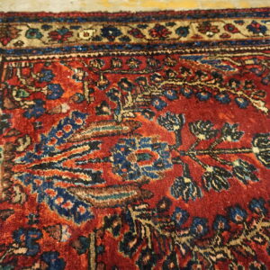 Textured, low-pile red rug with detailed traditional motifs in blue, tan, and black near the border.