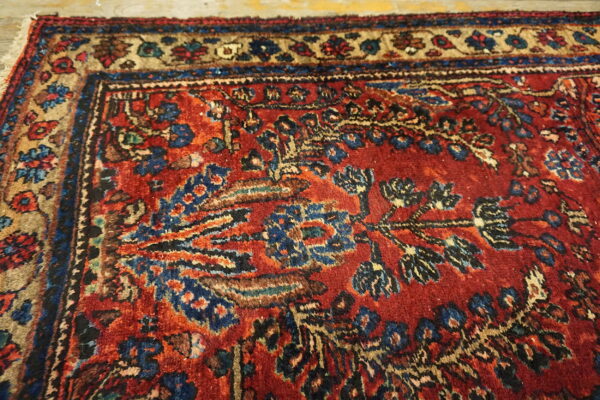Textured, low-pile red rug with detailed traditional motifs in blue, tan, and black near the border.