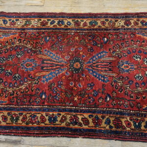 Distressed red runner rug featuring complex blue and tan floral patterns rests on light wood planks.