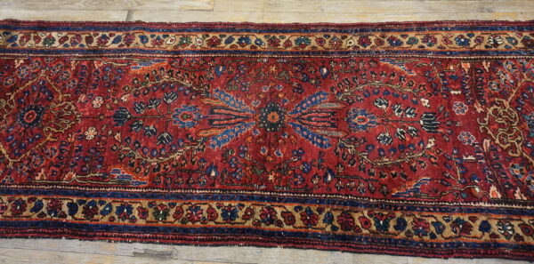 Distressed red runner rug featuring complex blue and tan floral patterns rests on light wood planks.