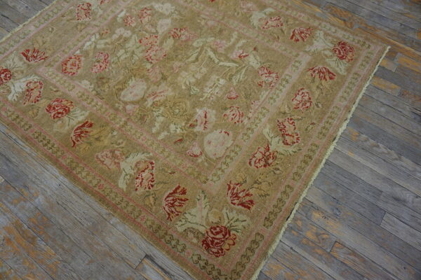 Low-pile tan floral rug with red and pink borders resting on rustic gray wood flooring.