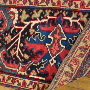 Red, navy, and blue geometric rug lying on hardwood, with a woven border corner folded back.