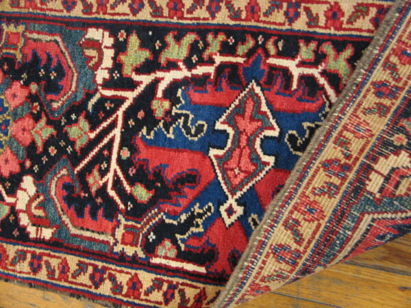 Red, navy, and blue geometric rug lying on hardwood, with a woven border corner folded back.