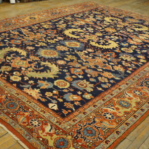 Low-pile rug featuring a navy blue field, intricate gold and rust floral motifs, and wide red borders on wood planks.