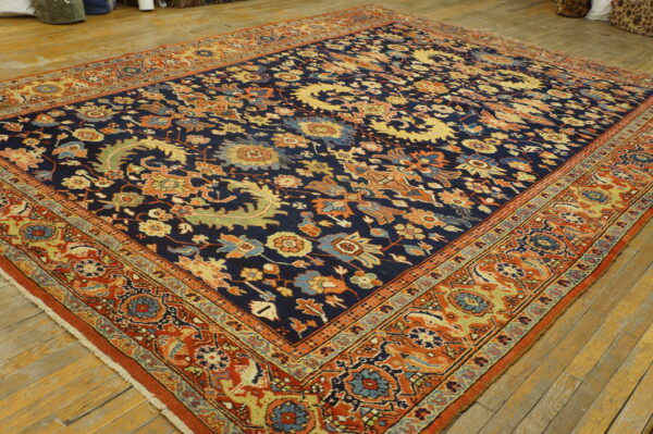 Low-pile rug featuring a navy blue field, intricate gold and rust floral motifs, and wide red borders on wood planks.
