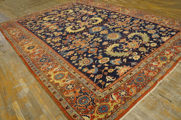 Large navy blue rug with dense floral motifs and a wide orange border rests on distressed hardwood floors.