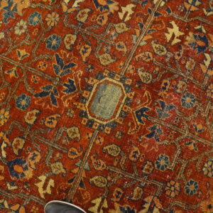 Heavily patterned red rug featuring faded blue and gold motifs, showing low pile texture and a shoe detail.
