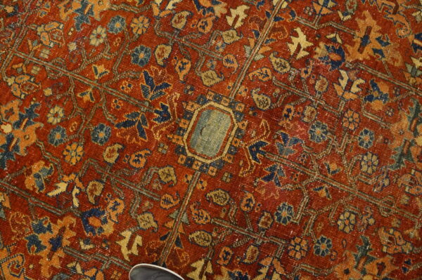 Heavily patterned red rug featuring faded blue and gold motifs, showing low pile texture and a shoe detail.