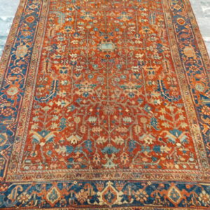 Traditional rug with a red field and blue border featuring detailed geometric patterns on a grey stone floor.