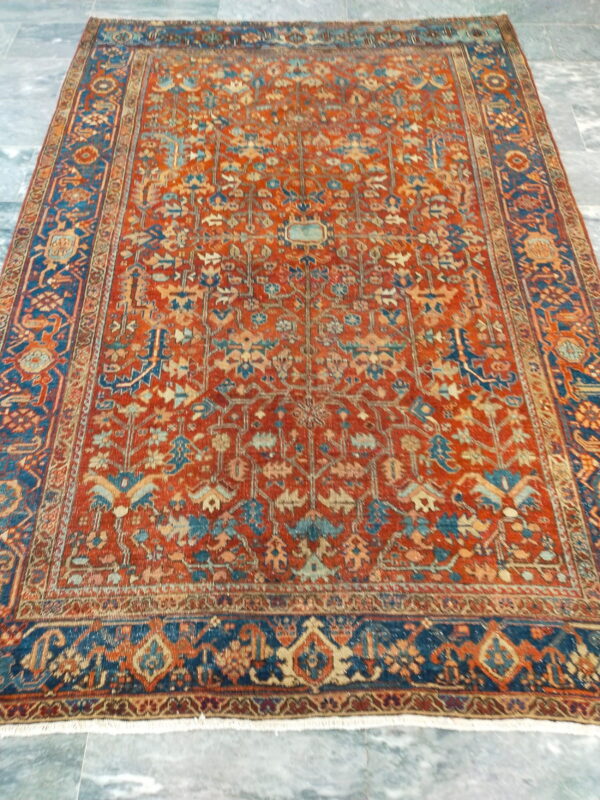 Traditional rug with a red field and blue border featuring detailed geometric patterns on a grey stone floor.