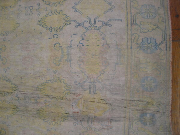 Distressed, low-pile rug close-up showing blue and yellow faded patterns and horizontal creases on wood floor.