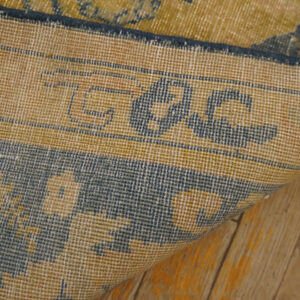 Tight weaving of a gold and dark blue patterned rug backing rests on distressed hardwood flooring.