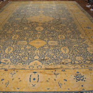 Ornate gold and blue rug spread flat on a wood floor near stacked rolled rugs.