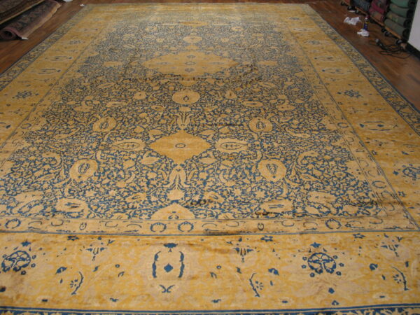 Ornate gold and blue rug spread flat on a wood floor near stacked rolled rugs.