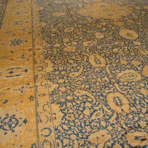 Detailed view of a golden yellow rug with dense blue scrolling floral patterns on dark wood flooring.