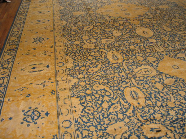 Detailed view of a golden yellow rug with dense blue scrolling floral patterns on dark wood flooring.