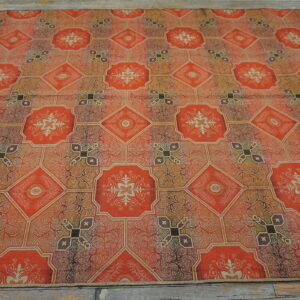 Orange and brown flatweave rug with repeating ornate medallions and geometric tiles covers a rustic wood floor.