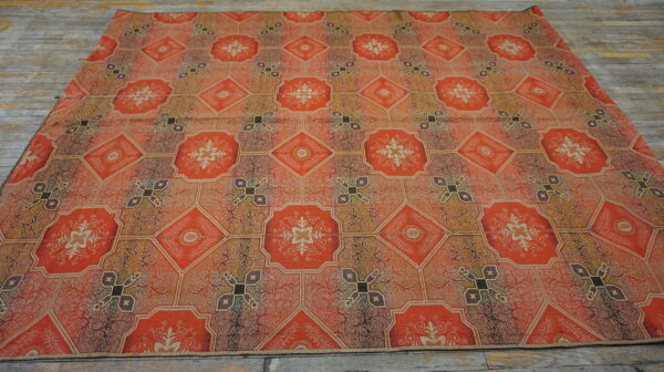 Orange and brown flatweave rug with repeating ornate medallions and geometric tiles covers a rustic wood floor.