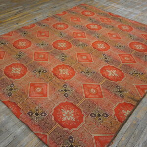 Ornate red-orange rug with repeating octagonal and diamond geometric patterns lies on worn wooden flooring.