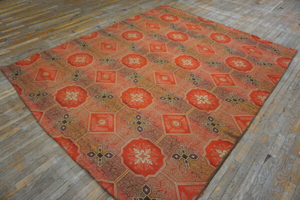 Ornate red-orange rug with repeating octagonal and diamond geometric patterns lies on worn wooden flooring.