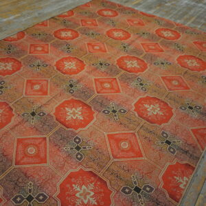 Intricate orange and brown tiled rug with floral and cross motifs displayed on rustic wood flooring.