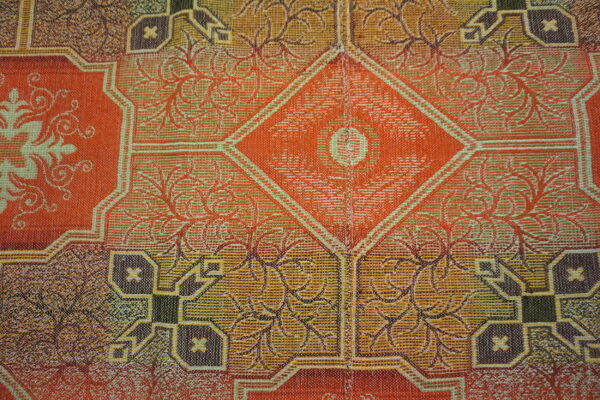 Detailed low-pile rug pattern featuring orange, gold, and brown geometric and branching floral designs.