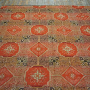 A rug featuring an orange, brown, and gold geometric tile pattern with floral medallions lies on weathered wood flooring.