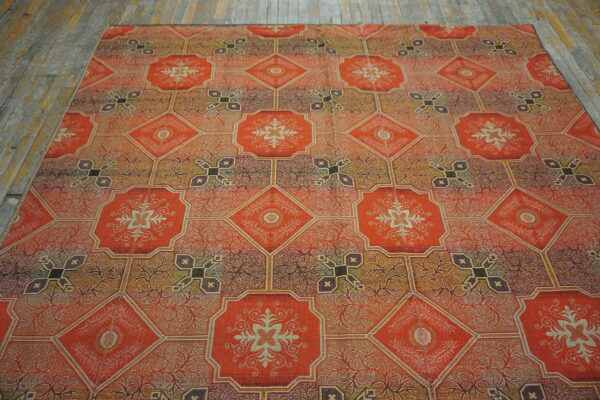A rug featuring an orange, brown, and gold geometric tile pattern with floral medallions lies on weathered wood flooring.