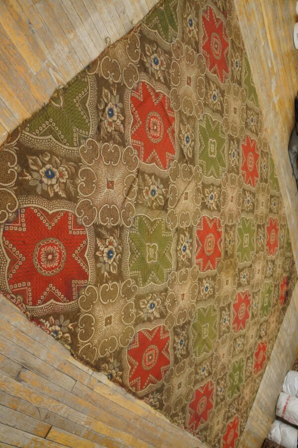 Brown, red, and olive green patterned textile featuring repeating star medallions on worn wood flooring.