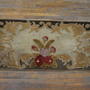 Low-pile beige rug with dark brown border, golden scrollwork, and central red and purple floral bouquet on wood flooring.