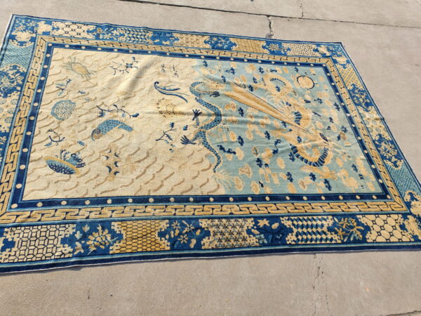 Blue and beige rug with a split field, central dragon, and geometric borders lies on concrete pavement.