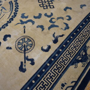 Tan rug with indigo traditional Chinese motifs and a geometric key border on worn wood planks.