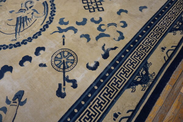 Tan rug with indigo traditional chinese motifs and a geometric key border on worn wood planks.